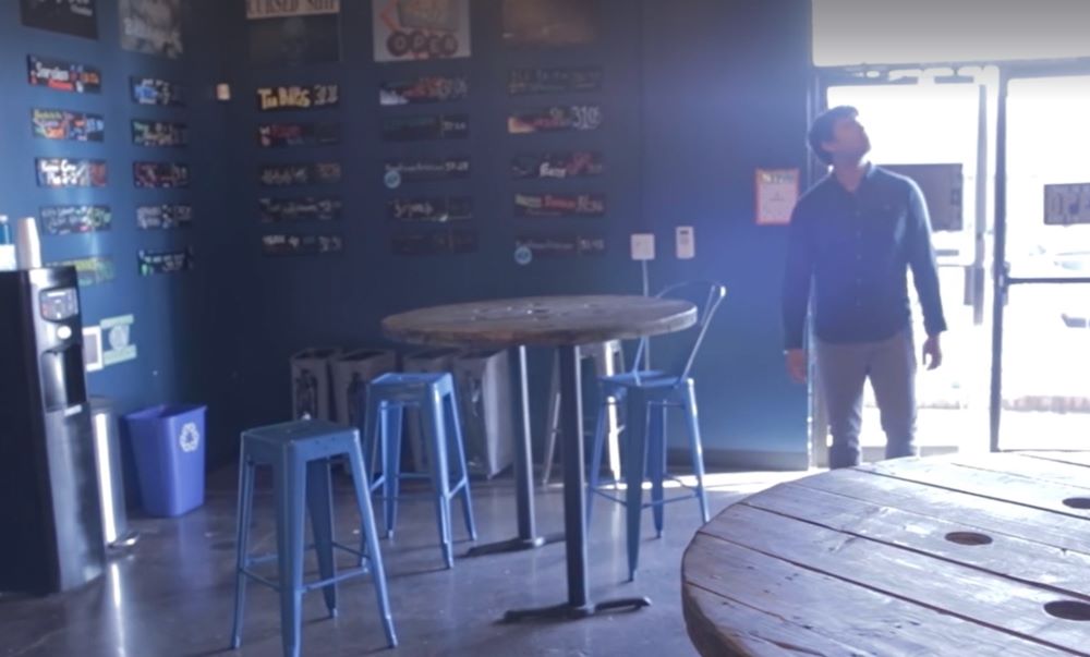 A screenshot from the movie "The Good Hearts Club" showing the main character Matt slowly stepping into the lobby of an escape room. Matt looks up at the ceiling as bright white light washes in through the glass doors behind him. To the left is a bar-height table with blue metal bar stools scattered around. The blue walls are display columns of decorated best times for each of the escape rooms in the business.