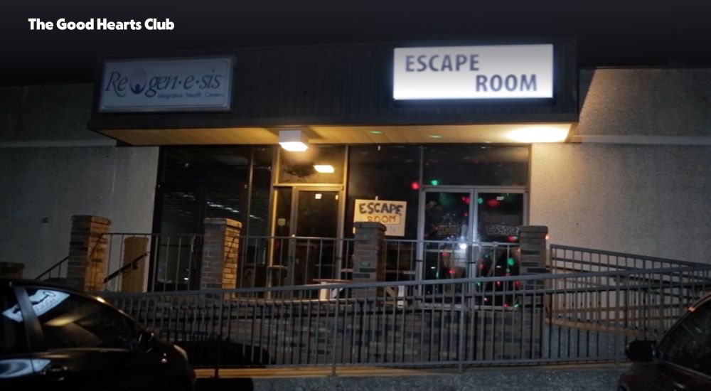 A screenshot from the movie "The Good Hearts Club" shows the storefront for a simple, no frills escape room. It's dark outside, so our eyes are drawn to the barebones "Escape Room" sign lit above tthe doorway.
