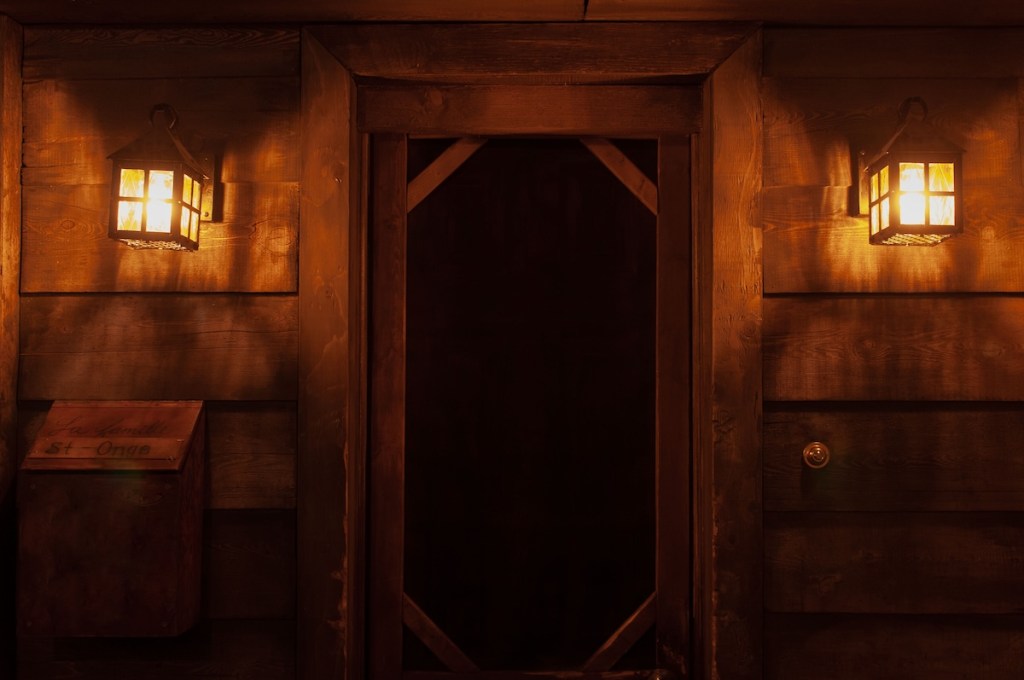 The front door to an old, ominous wood cabin. The door is flanked by two illuminated lights.
