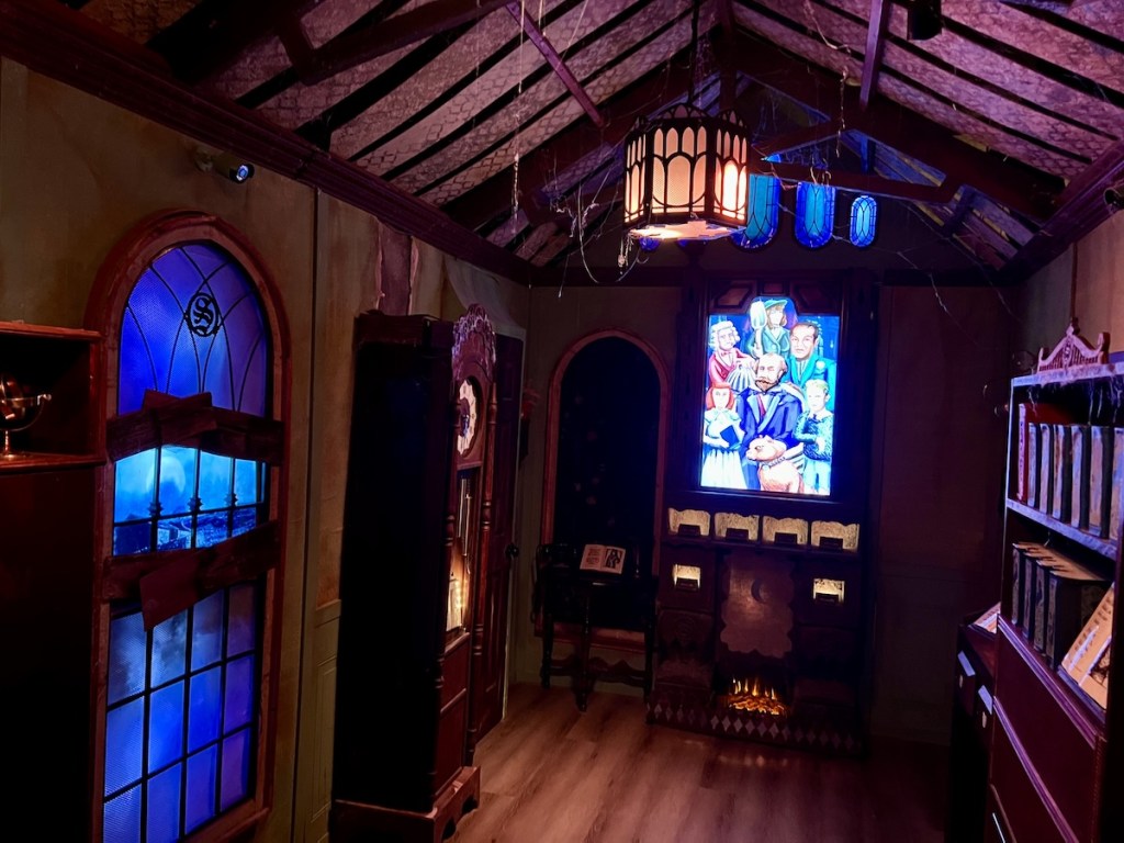 A long room in an old, rundown mansion with a bookcase, a grandfather clock, a family portrait, and stained glass wndows.