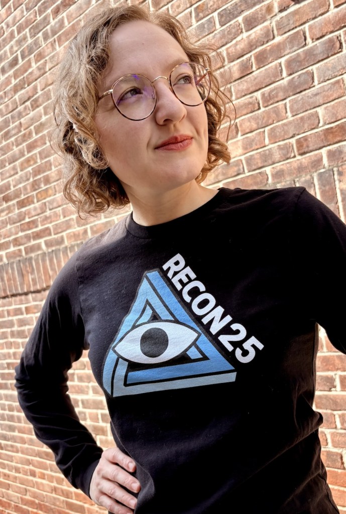 Blonde woman in glasses wearing a black RECON 25 shirt.