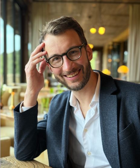 Man smiling in grey suit, white button down shirt with glasses and beard