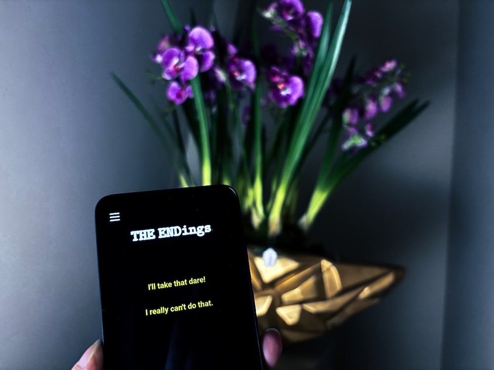 A mobile device that reads, "The ENDings," yellow text below reads "I'll take that dare!" and "I really can't do that." Beyond the device is a large orchid arrangement in a gold vase.