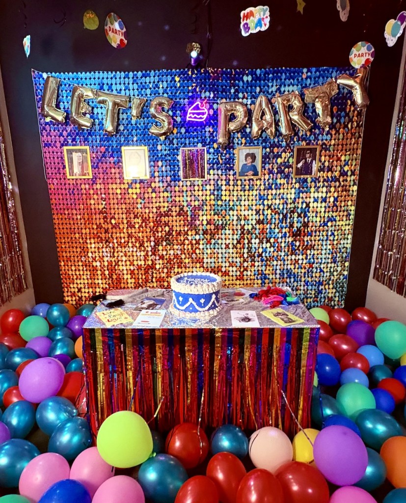 A party room with balloons covering the floor, a table with a cake, and an iridescent wall that has a sign that reads, "Let's Party."