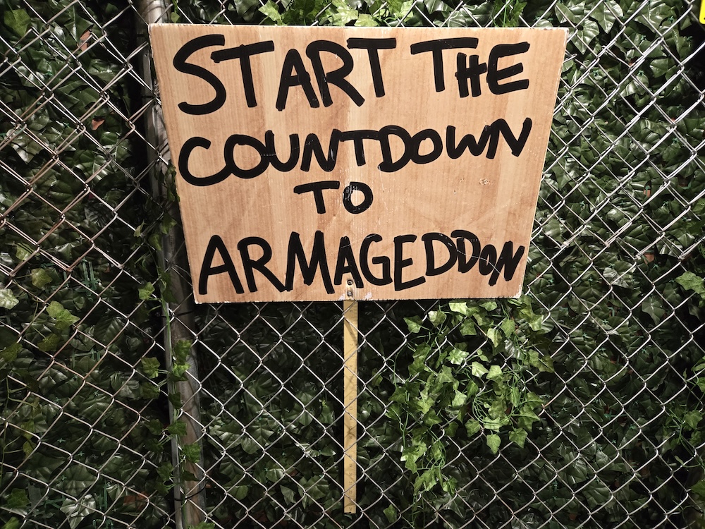 A hand painted sign stuck in a chainlink fence reads, "START THE COUNTDOWN TO ARMAGEDDON"