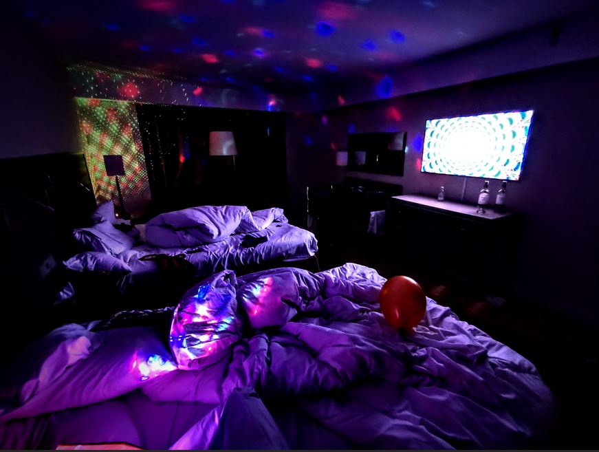 a dark hotel room lit only by disco lights and graphics playing on the tv. there are balloons and bottles of alcohol laying around.