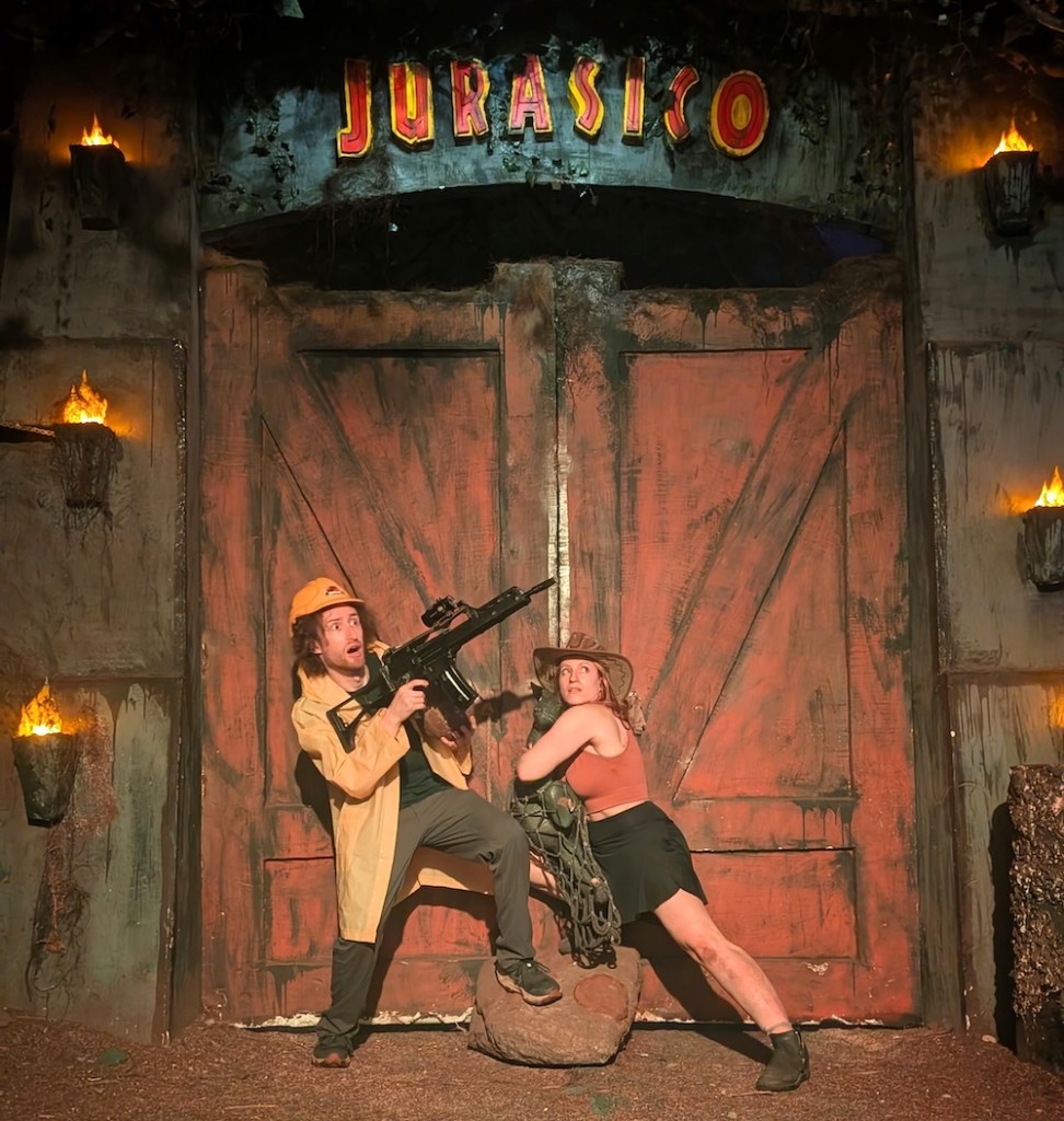 A man holding an assault rifle, a woman holding a baby dinosaur, all in front of a large wooden gateway labeled, "JURASICO"