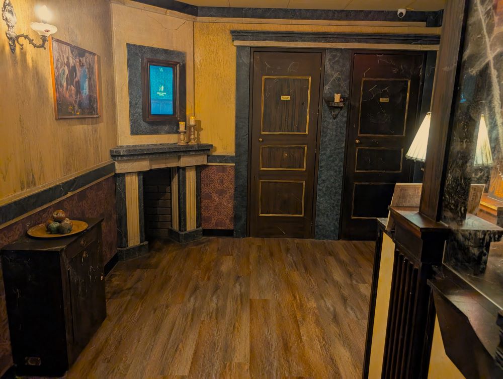 The warmly lit lobby of a hotel as viewed from in front of the reception desk. Across the room are two dark wooden doors framed by dark grey stone. To the left of the doors is a fireplace in the corner with a framed display screen hanging on the wall above it.