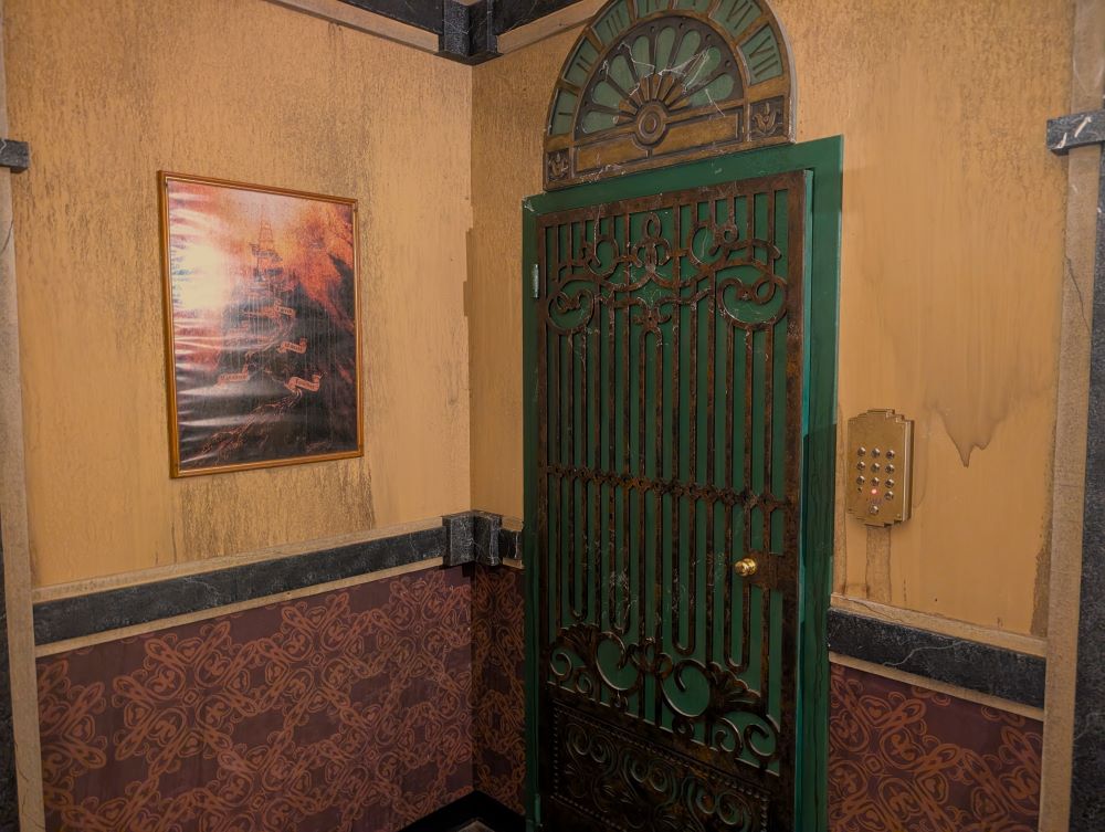 A green and bronze-grated door masquerades as an old-timey elevator in the corner of a hotel lobby. Above the door, a green and bronze arch immitates the floor indicator of the elevator, showing Roman numerals from 1 to 7 along the arc of the arch.