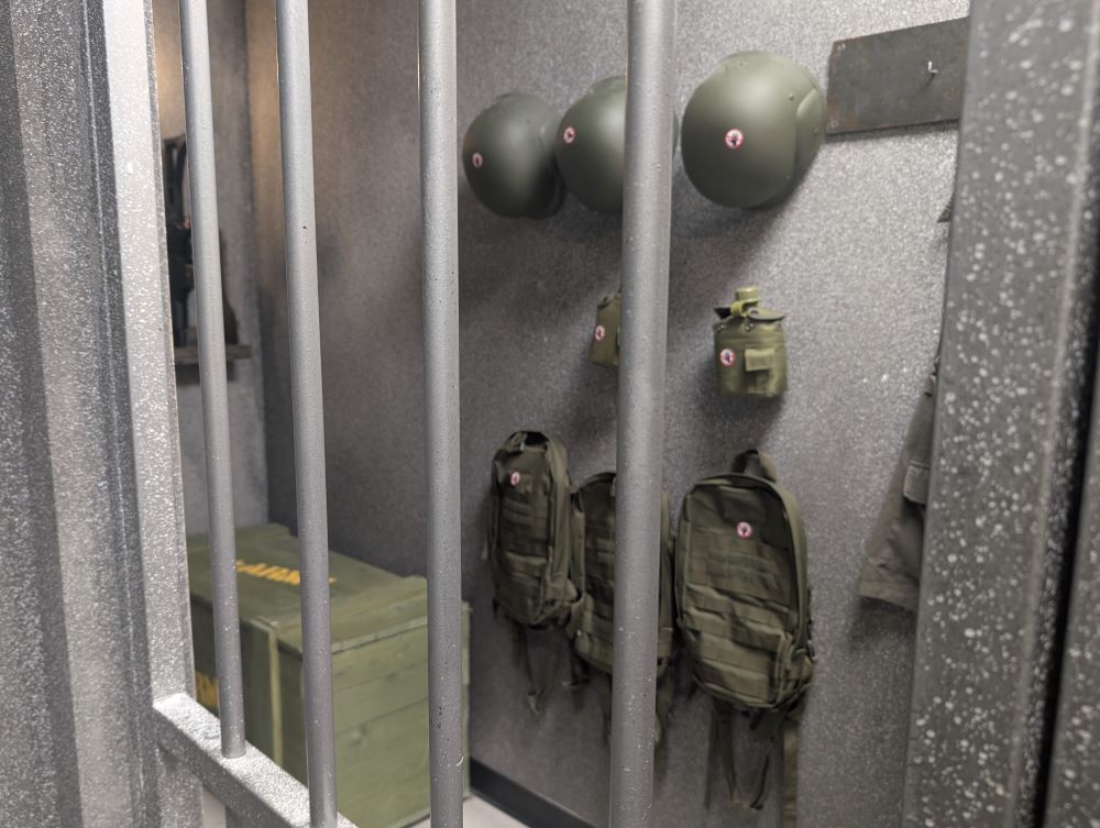 A view through a barred prison cell door showing a set of army helmets, canteens, and backpacks hanging on the wall out of reach.