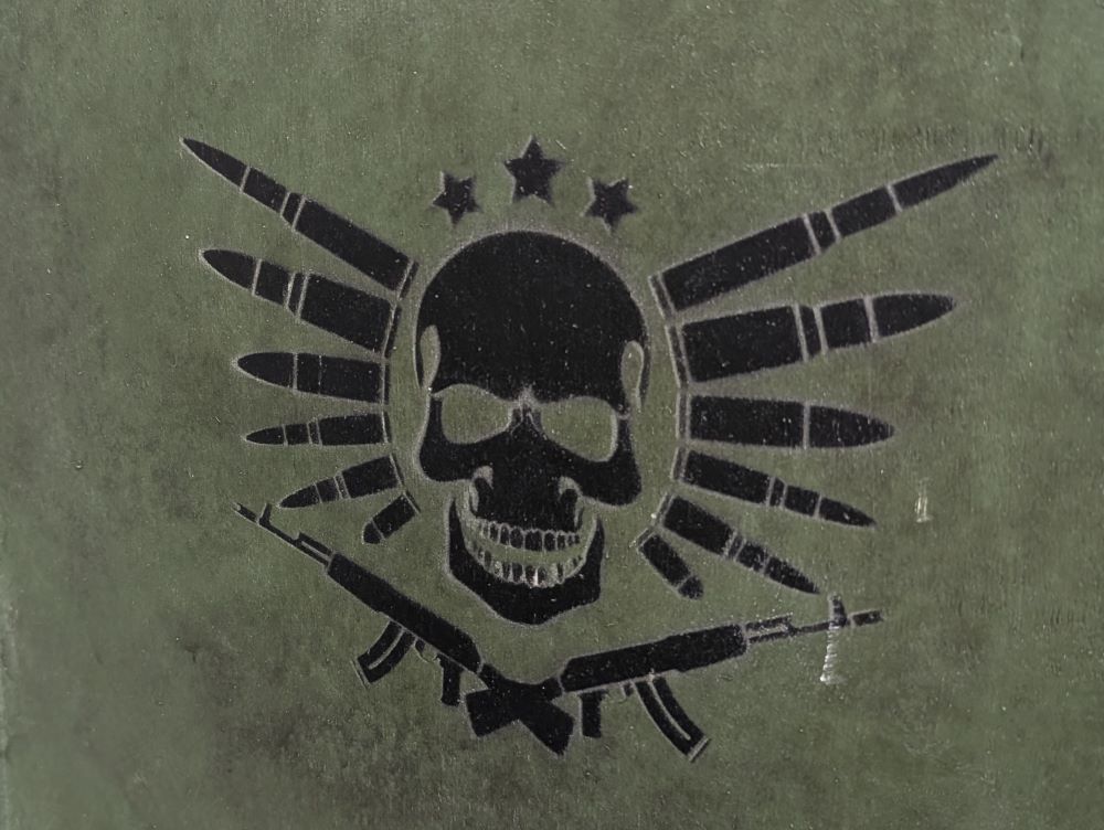 A black logo printed on an army-green background showing a skull in the center, three stars in an arc above, a "wing" of six bullets on either side, and two machine guns crossed below.