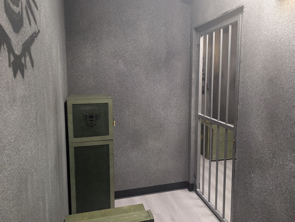 A small, sparse grey cell. A locked greenish cabinet stands in the far left corner. A barred cell door is located in the far right corner.