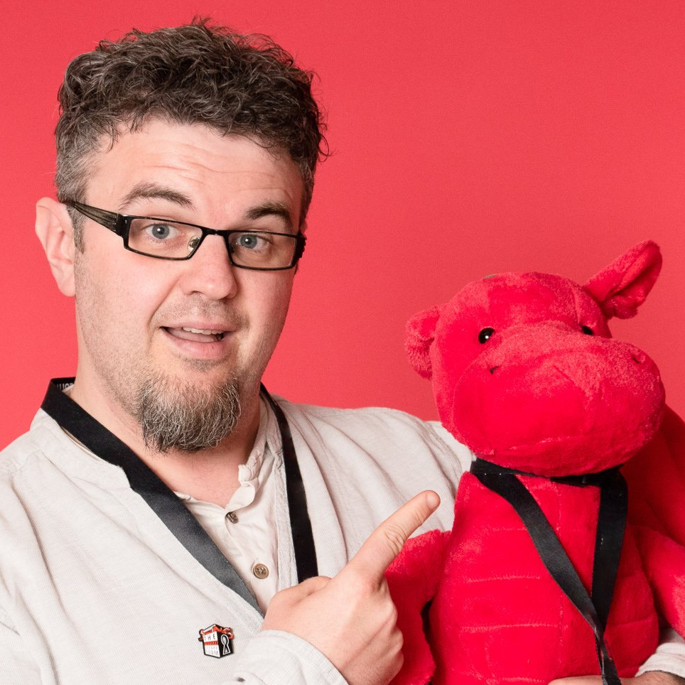 man with wavy brown hair and grey eyes with glasses and a goatee. he is wearing an ecru long sleeve shirt and holding a red hippo.