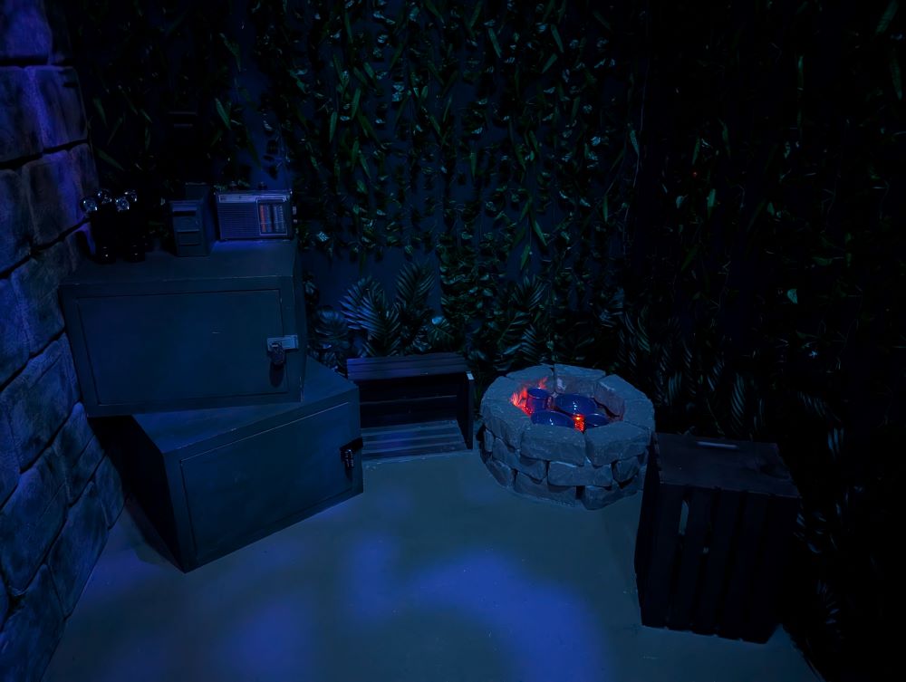 Photo of an explorer's campsite. A small fire pit is lit and surrounded by locked crates and foliage.