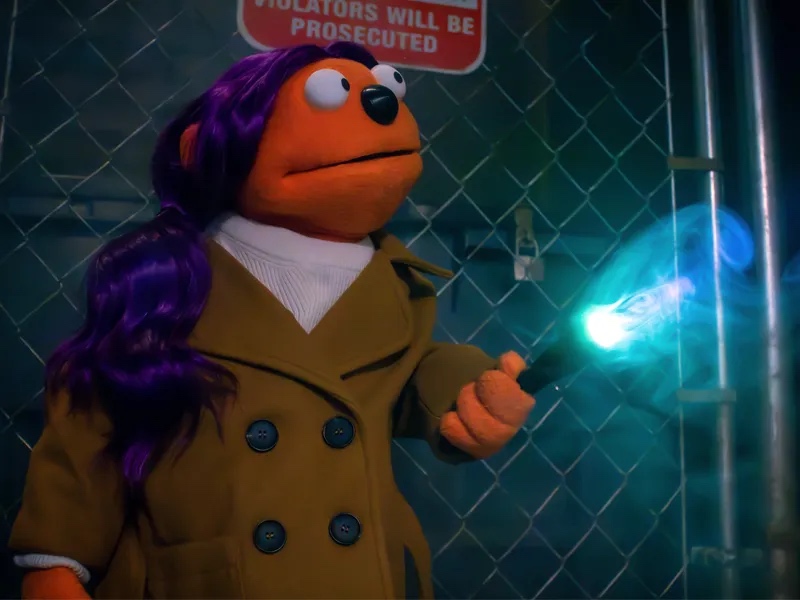 A purple-haired orange puppet investigating something with a flashlight.