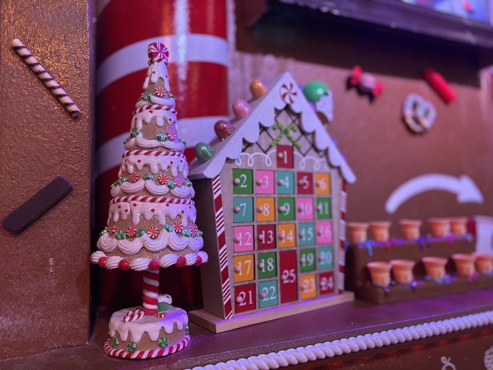 Various sweet Christmas decorations on a fireplace mantle. A frosted gingerbread tree is next to a colorful house-shaped advent calendar.