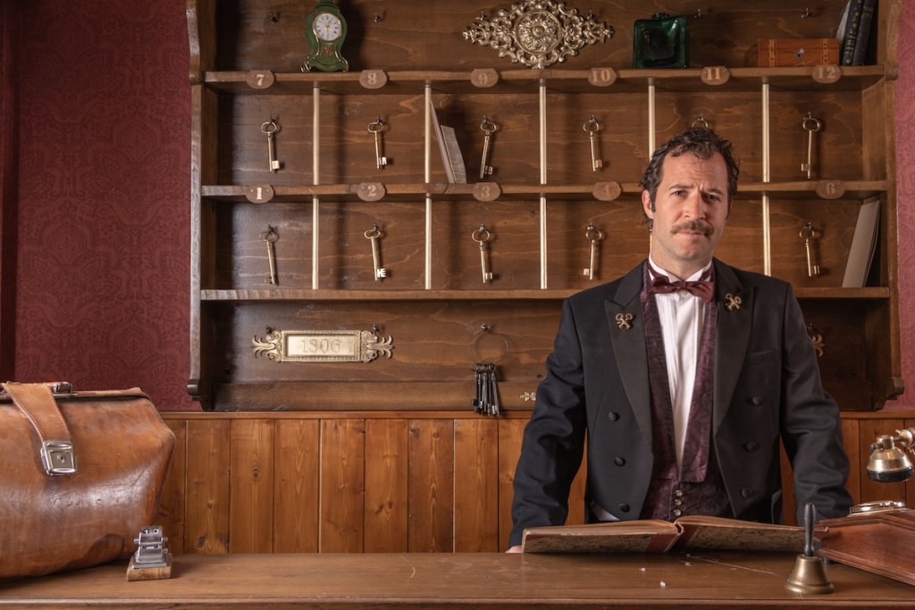 The front desk of a fancy hotel. A well-dressed concierge stands in front of a wall of hanging keys.