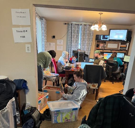 2 people sorting through flattened cereal boxes, surrounded by large plastic bins filled with flattened cereal boxes, and people on computers in the background. paper taped to the walls with trivia ID # and phone numbers