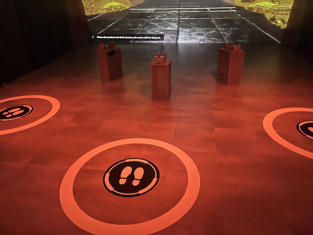 Three small circles with shoeprint icons are on the floor, telling players where to stand for proper motion detection. There is a small grey pedestal for each player to set their device in. The very bottom of a scene projected onto the front wall can be seen, showing mountains of gold items to each side.