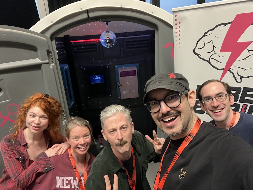 David and friends taking a selfie in front of a porta-potty that has been turned into an escape room with a disco ball.