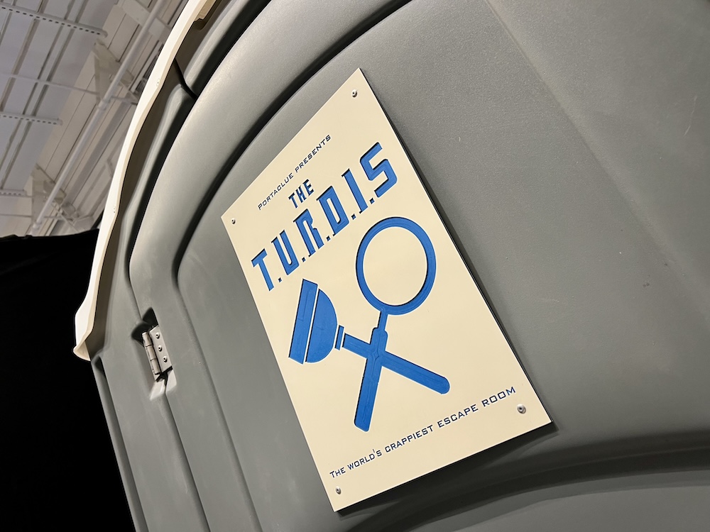 A sign on the front of a porta-potty reads, "The TURDIS, the World's Crappiest Escape Room"