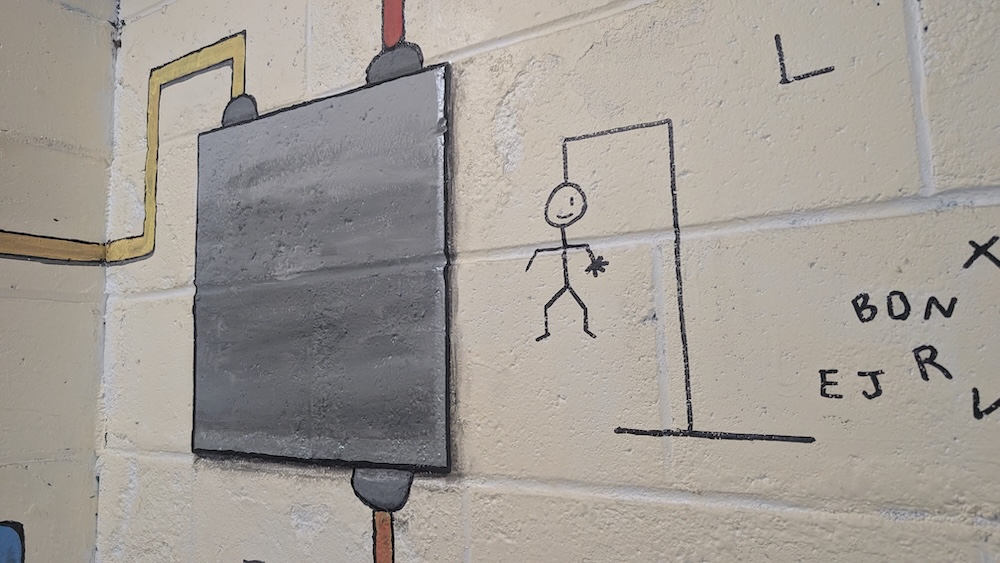 Puzzle mural painted on a concrete wall depicting a junction box and a partial hangman game beside assorted letters.