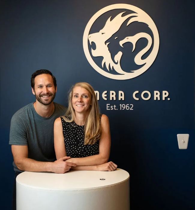 man smiling with very short beard and side parted neat hair, wearing a grey t-shirt. posed holding hands with a woman with long blond hair, smiling, in a polka dotted sleeveless top.