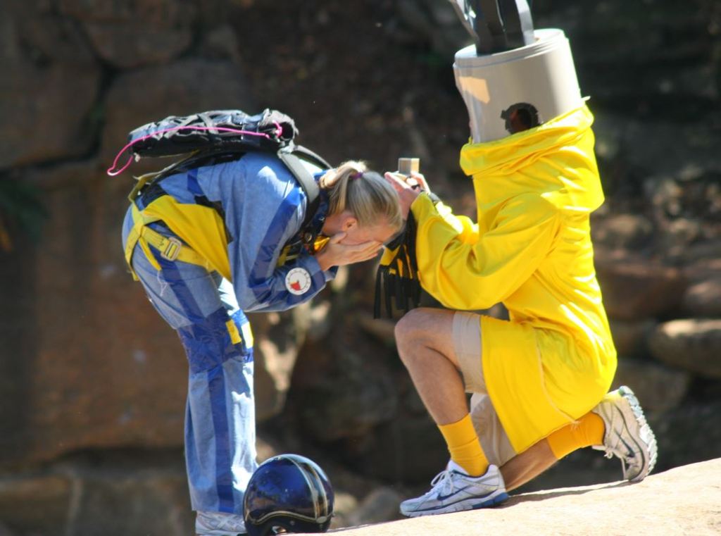 blonde woman in blue skydiving jumpsuit and helmet bending over covering her face. man in yellow air dancer costume on one knee, holding an engagement ring box.