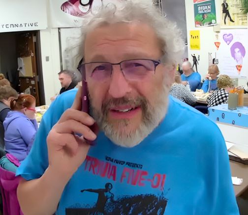 man with shaggy grey hair and a white beard, wearing glasses and a bright blue t-shirt. He is on the phone, standing in a room full of people answering phones.