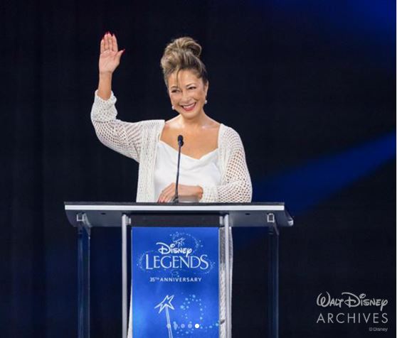 eurasian woman standing at podium that reads "disney legends", she is wearing white and waving