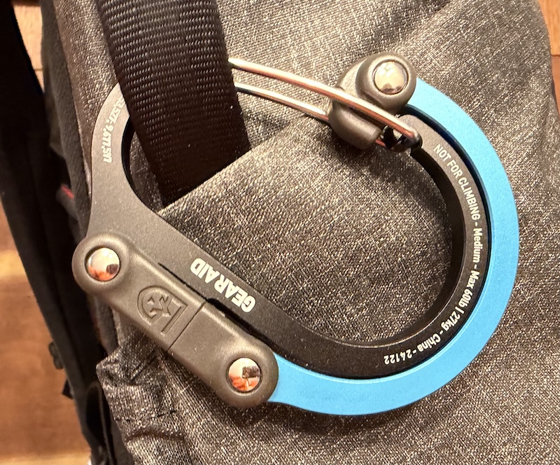 An elaborate carabiner with an extra flex-point hooked around the handle of a backpack.