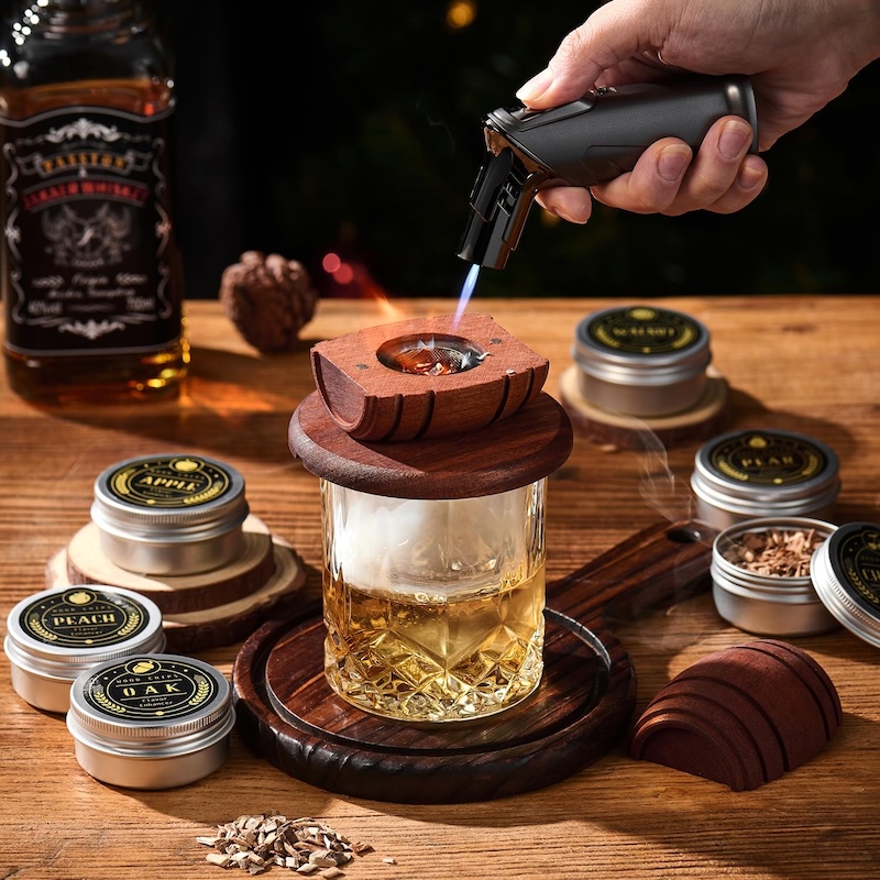 A whiskey smoking apparatus sitting atop a glass of whiskey. A small blow torch toasts wood chips to provide the smoke.