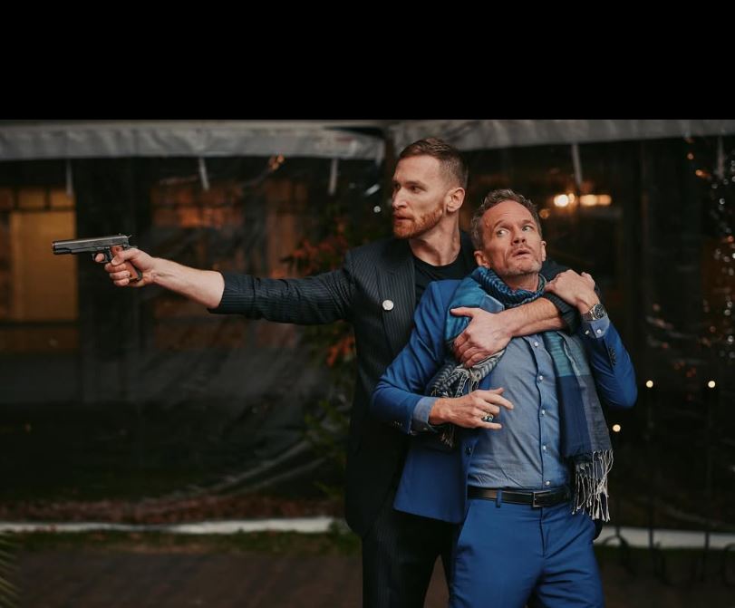 man with short red beard in pinstriped suit and black t-shirt is holding a gun outward and the other arm is wrapped around a man (NPH) in a blue suit and blue patterned scarf, like he is holding him hostage