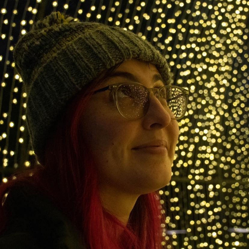 woman wearing glasses and green beanie with pink hair, looking up and surrounded by twinkling christmas lights