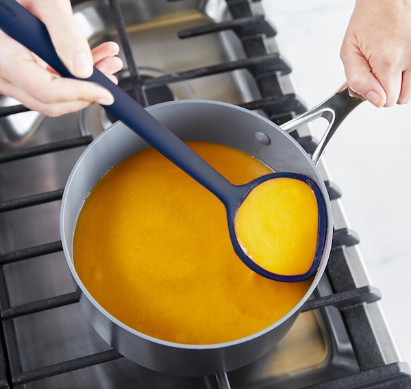 A ladle with a flat, flexible front scooping soup from a pot.