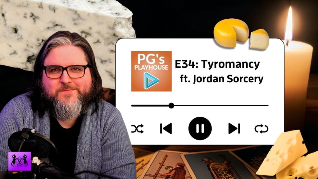 man with white streak in dark brown, shoulder length hair, brown beard with white streak, wearing glasses and grey cardigan. background has candle, tarot cards, and various cheeses