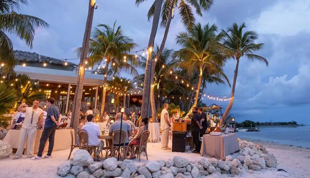 outdoor dinner on the beach with palm trees and people sitting at dining tables with an outdoor bar. there's lights strung up and a low rock wall surrounding the dining area