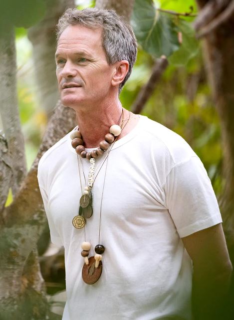 man with greyish brown hair in white t-shirt, wearing several necklaces with large wooden beads, standing in a jungle.