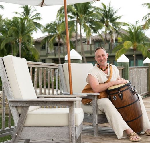 man dressed in loose white clothing, seated, playing large bongo in a tropical resort with palm trees in the back