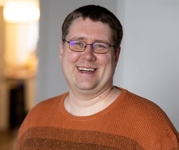 man with short brown hair, glasses, blue eyes, smiling joyfully, wearing a dark orange sweater