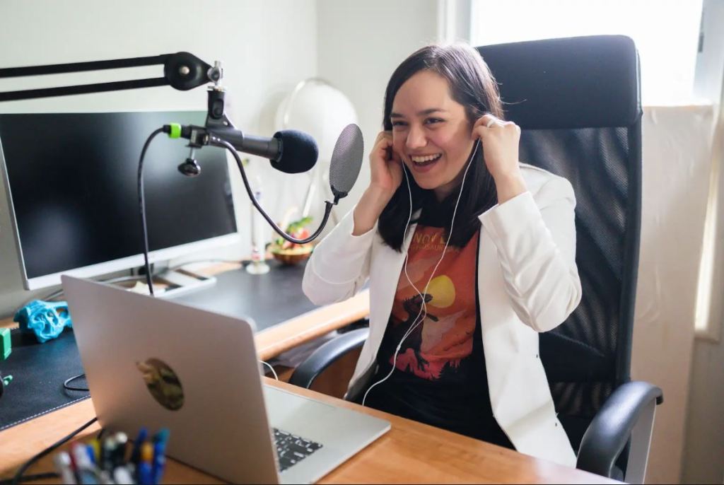 dark haired woman in a white blazer and black t-shirt sitting at a podcast studio desk