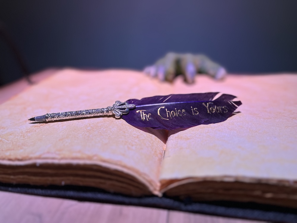 An open book with a quill pen. The feather of the quill reads, "The Choice is Yours" in gold lettering.