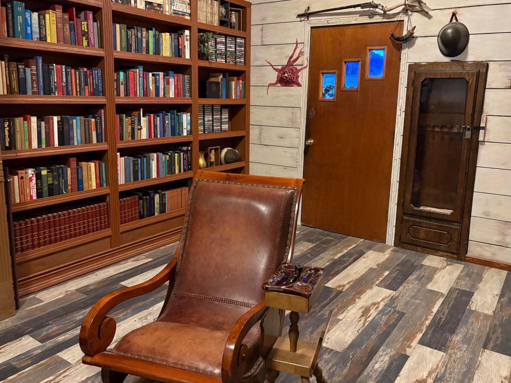 Room view with a leather chair and table in the center. A large bookcase filled with books sits along one wall, while a door with 3 windows diagonally shows a view of the lake beyond.