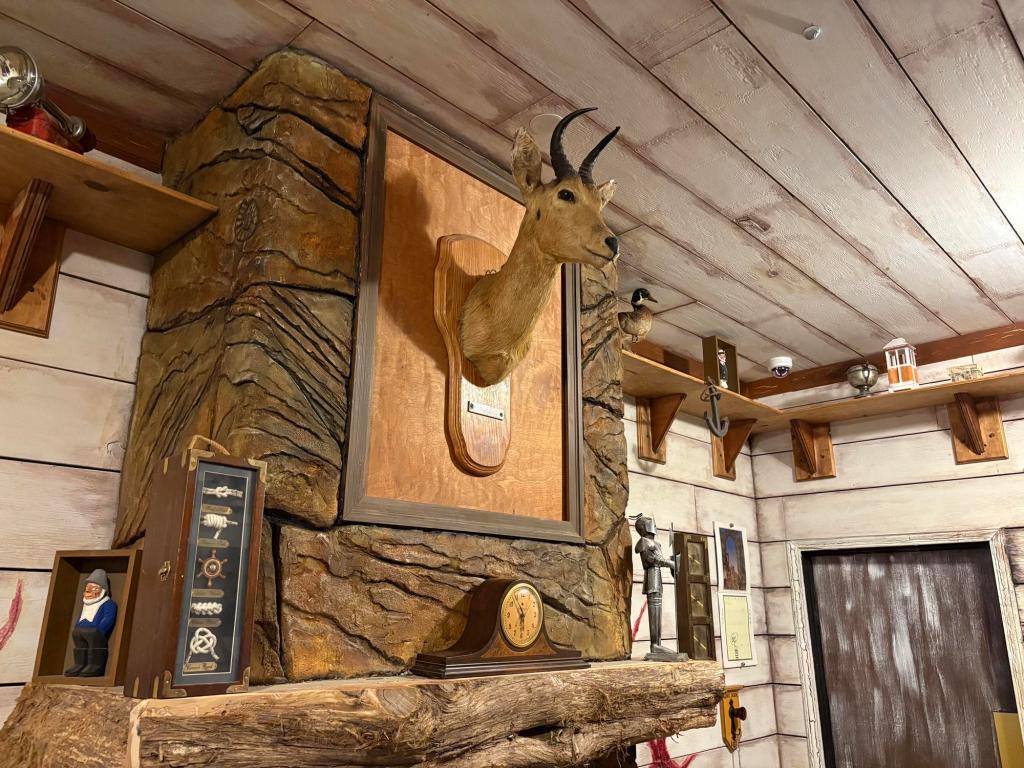 A deer head is mounted on a stone fireplace overmantel, various nautical decor sits on the fireplace mantel. The ceiling is made of wooden planks painted white.