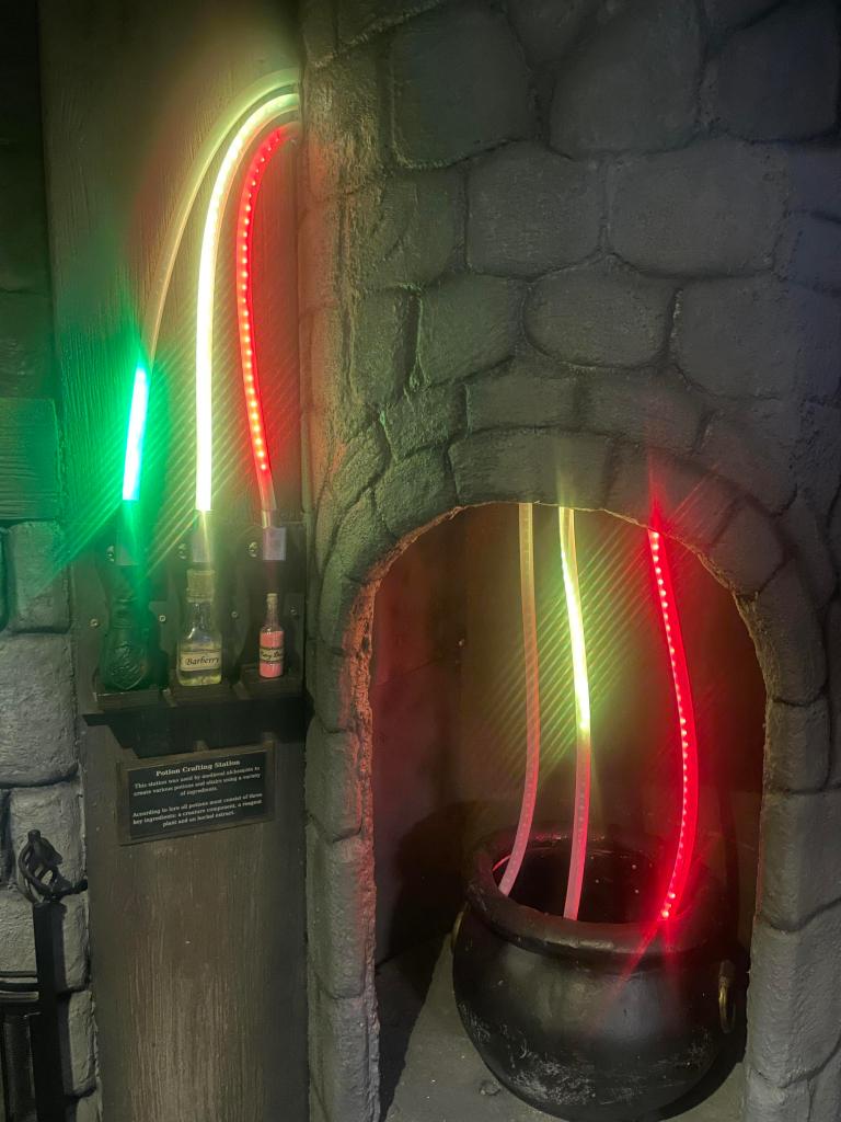 A cauldron sits within a stone fire place and chimney. LED light tubes flow up from the cauldron through the stone structure and connect to bottles on a shelf next to it.