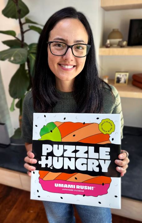 woman smiling with long straight black hair with black glasses. Wearing green grey short sleeve sweater and holding a game called puzzle hungry