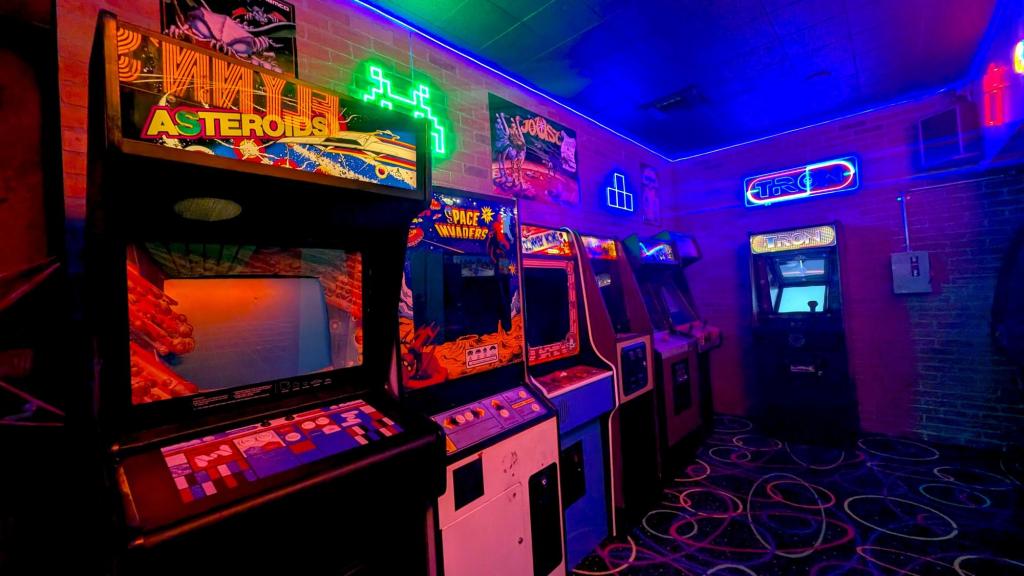 Seven arcade machines are in a room with neon signs and posters on the brick wall. The carpet is covered in a neon 80s style.