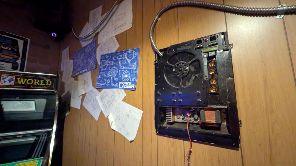 A wood paneled wall shows a mysterious electronic board. There also are numerous printouts and machine blueprints tacked to the wall.