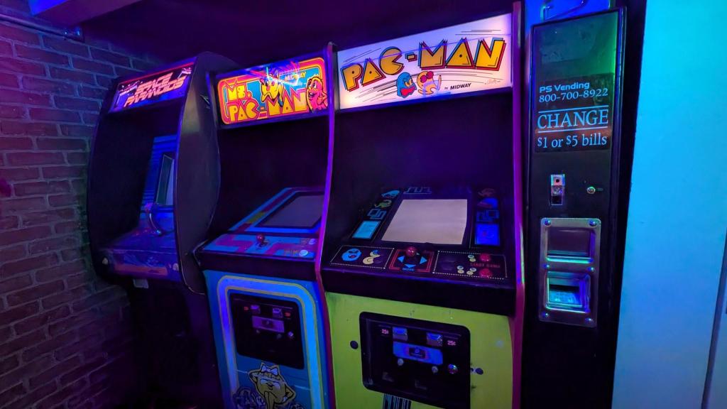 Three arcade machines and a coin changing machine are shown in a corner of a brick-walled room.