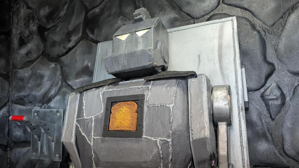 A gray robot against a gray stone while. The robot has a piece of bread front and center on its chest. There is a red lever to its right.